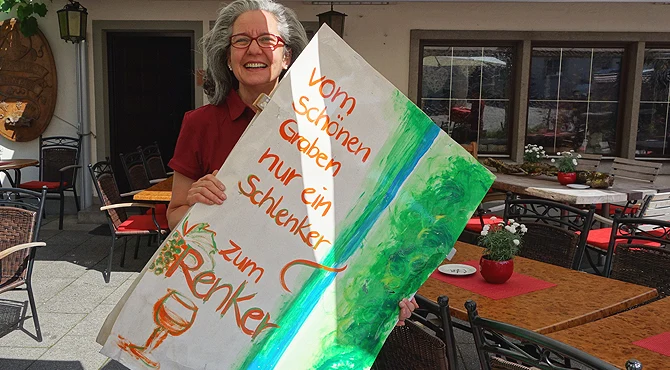Elke Renker lachend mit einem handgemalten Schild vor ihrer Weinstube
